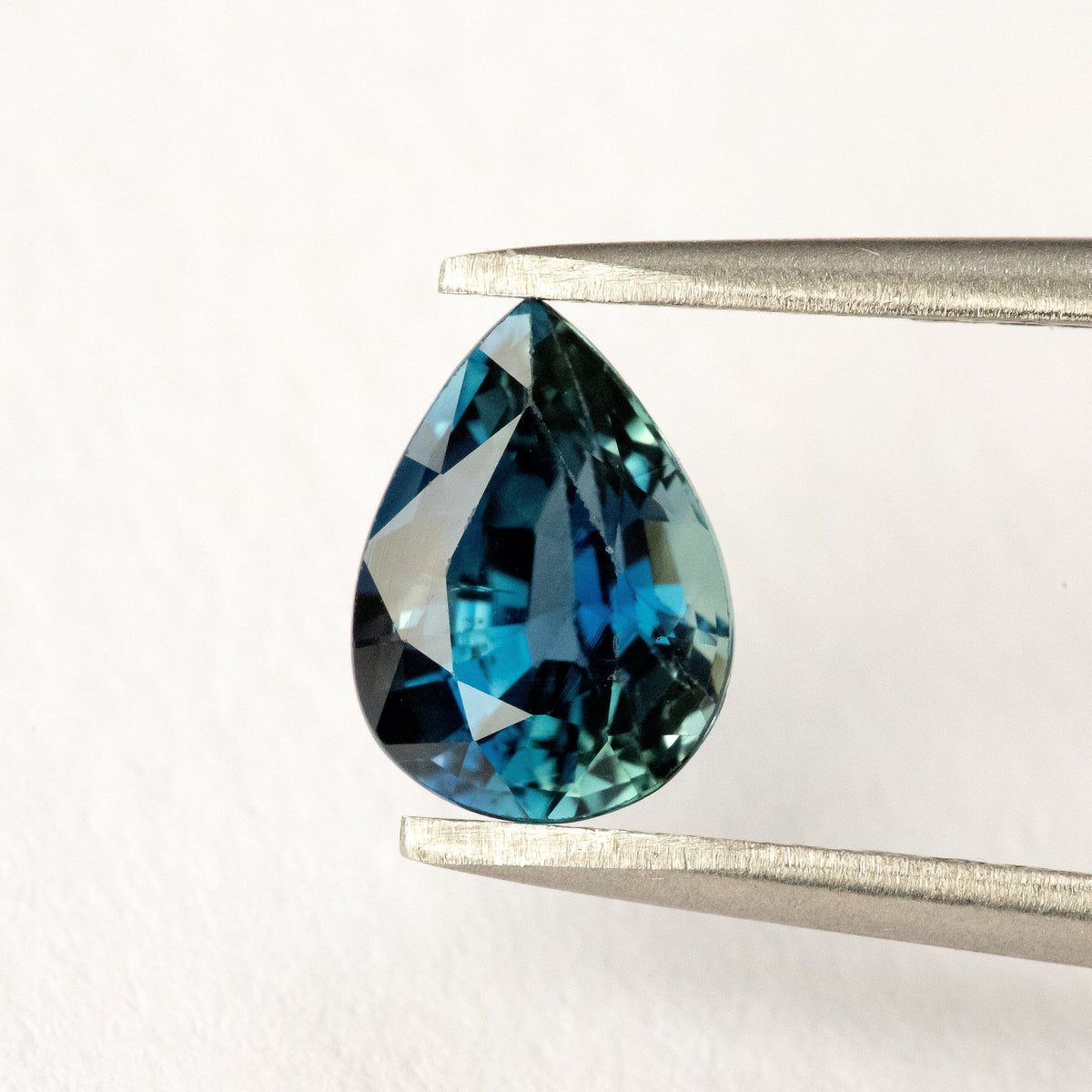 Blue-green pear-shaped sapphire held upright in tweezers, with dark central facets and bright blue reflections near the rounded end.