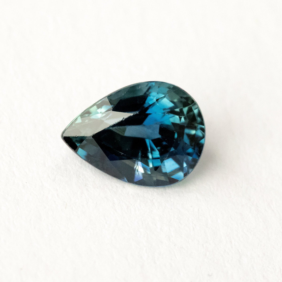Pear-shaped blue-green sapphire lying sideways on a white surface, showing dark triangular crown reflections.