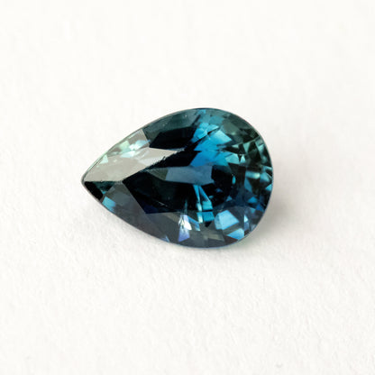 Pear-shaped blue-green sapphire lying sideways on a white surface, showing dark triangular crown reflections.