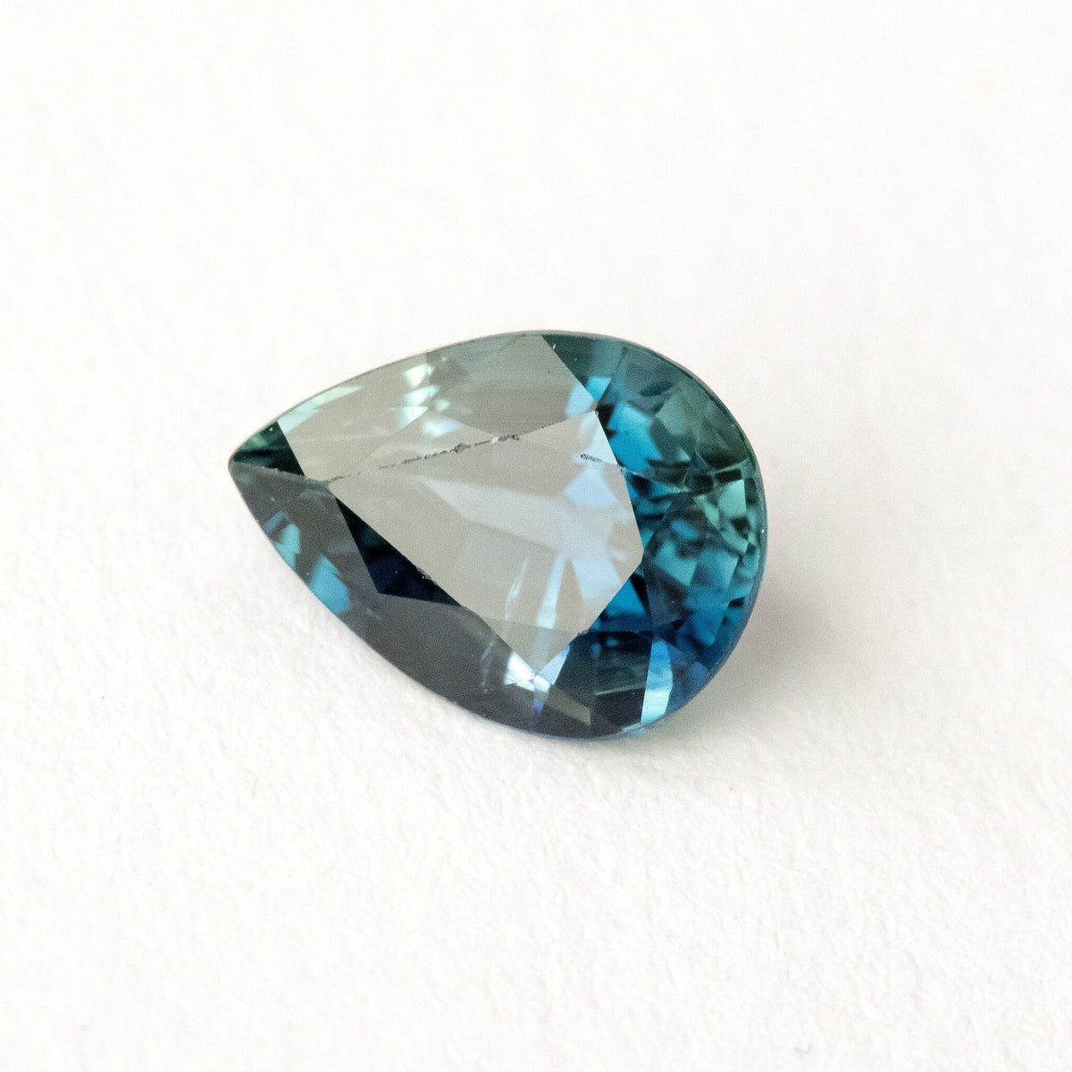 Blue-green pear-shaped sapphire on white, tilted to show a broad pale reflection across the table.