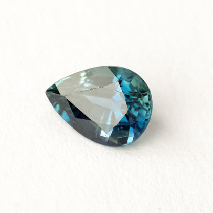 Blue-green pear-shaped sapphire on white, tilted to show a broad pale reflection across the table.