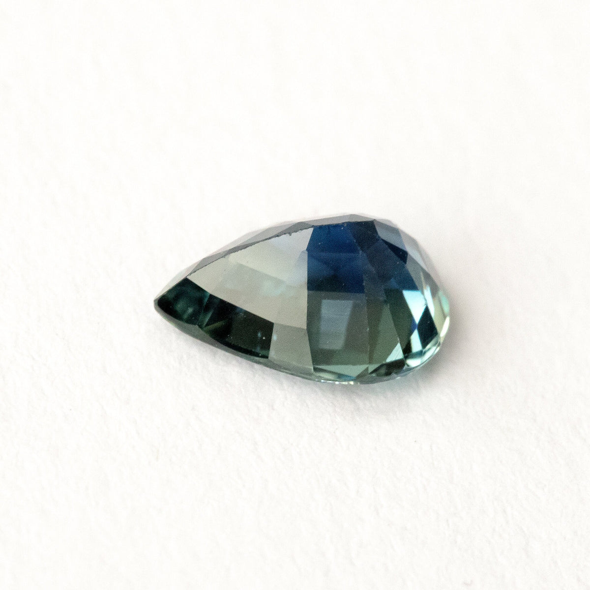 Pale blue-green pavilion of the pear-shaped sapphire resting on white at an oblique angle.