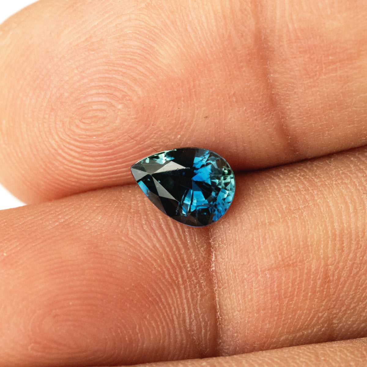 Blue-green pear-shaped sapphire resting on two fingertips, with its point facing left and blue flashes near the rounded end.