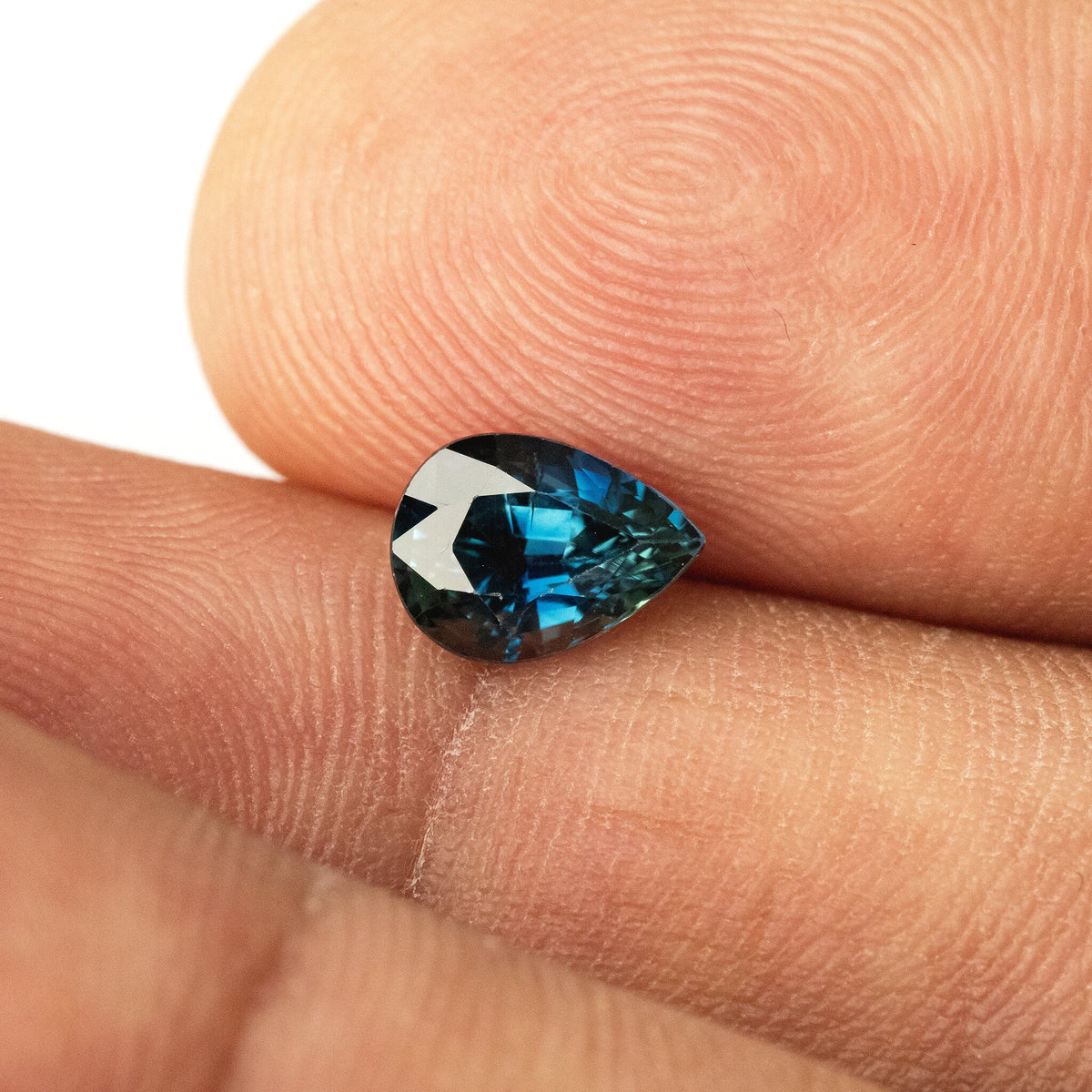 Pear-shaped sapphire against fingertips, showing its small scale and dark blue-green face-up reflections.