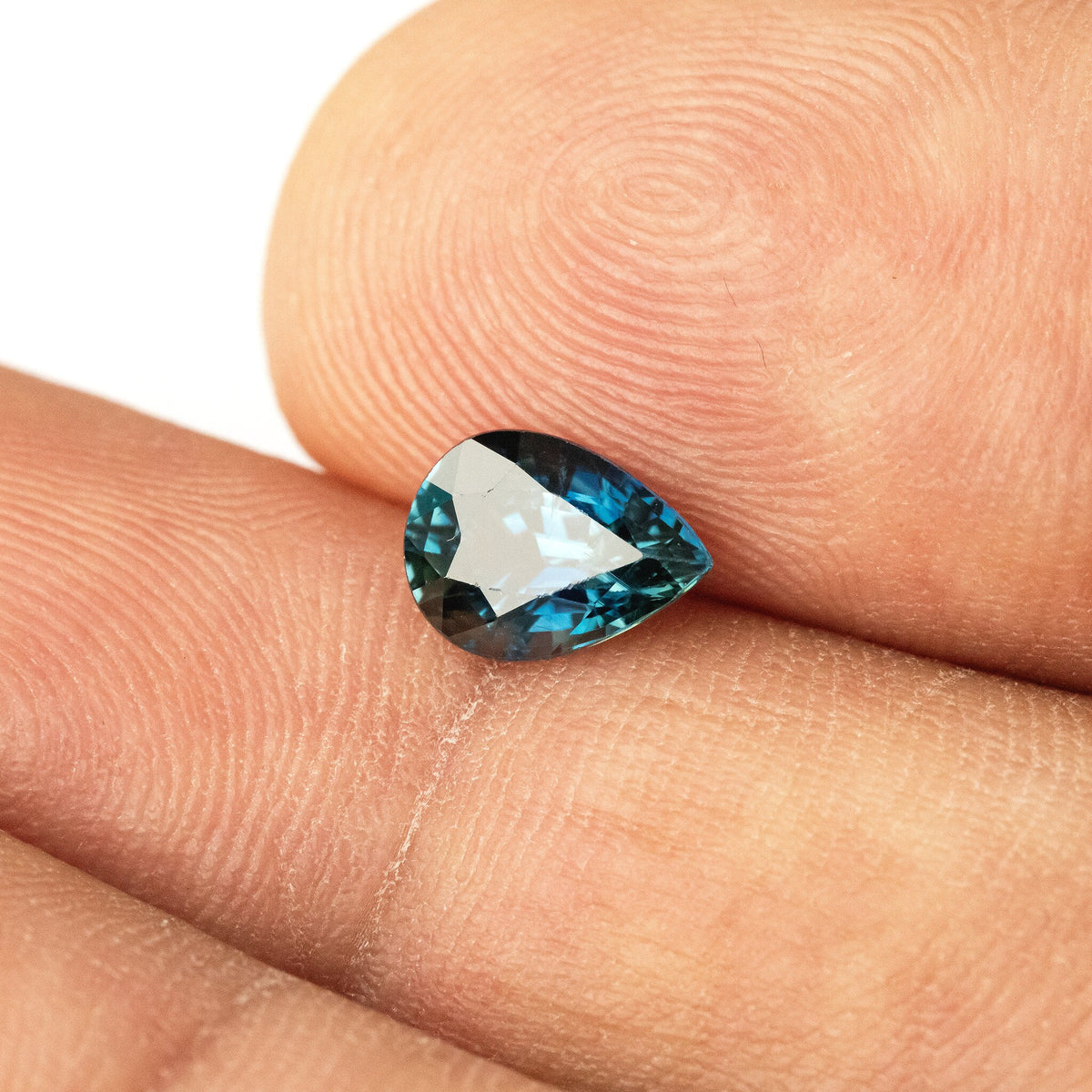 Pear-shaped sapphire between fingertips with a bright central reflection and blue facets around the edge.