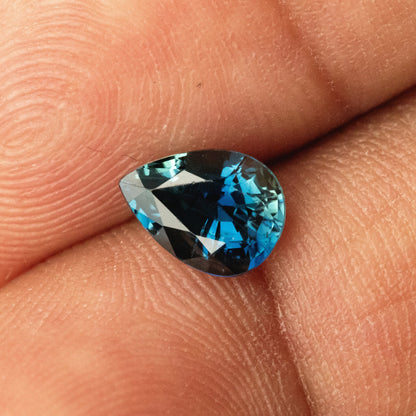 Close view of the pear-shaped sapphire on a finger, with its pointed left tip and blue-green facet reflections visible.