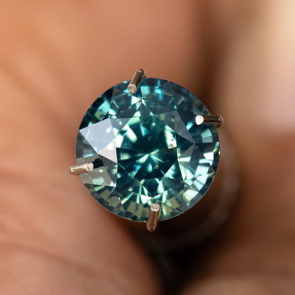 Round teal-blue and green sapphire at a slight face-up tilt in a temporary prong-style inspection holder; holder not included.