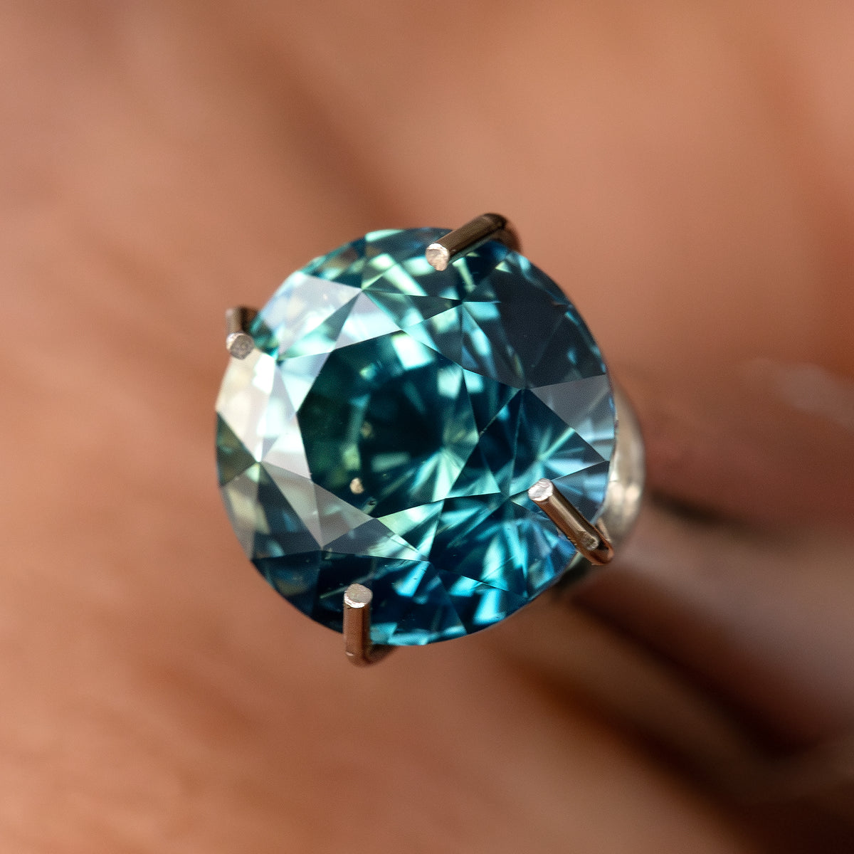 Oblique face-up view of the round teal-blue and green sapphire in a temporary prong-style inspection holder; holder not included.