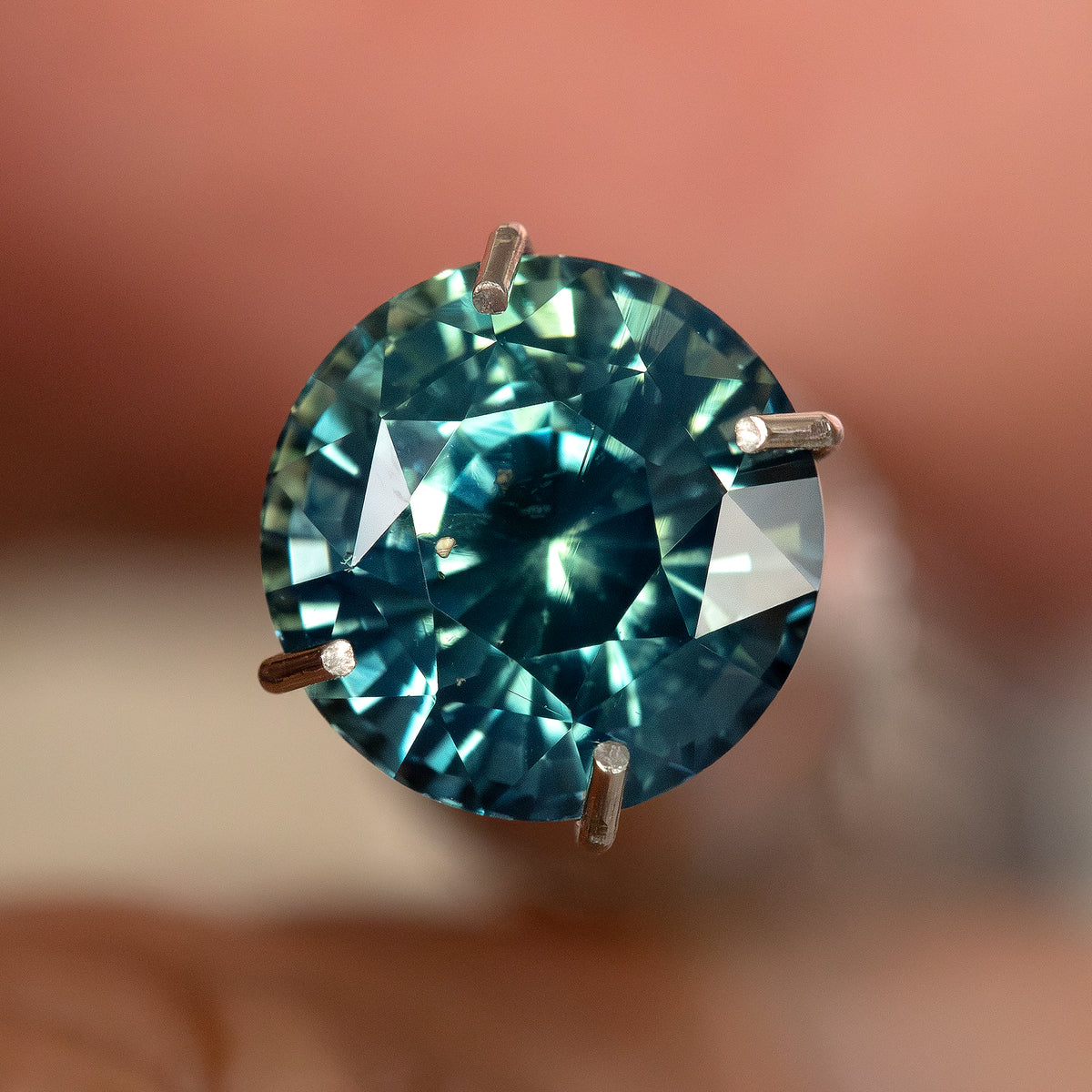 Round teal-blue and green sapphire face-up in a temporary prong-style inspection holder, with blue-green flashes; holder not included.