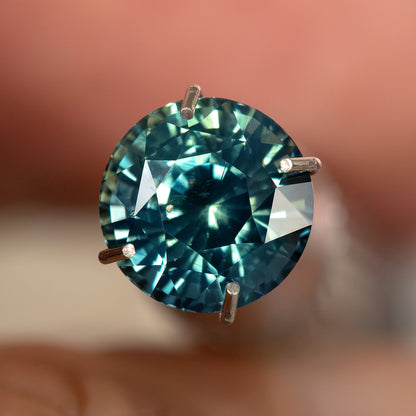Round teal-blue and green sapphire face-up in a temporary prong-style inspection holder, with blue-green flashes; holder not included.