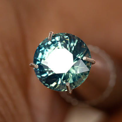 Round teal-blue and green sapphire in a temporary prong-style inspection holder, with a large white table reflection; holder not included.