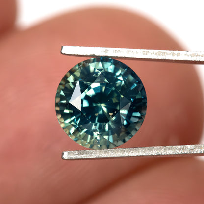 Round teal-blue and green sapphire face-up in tweezers against a fingertip for scale.