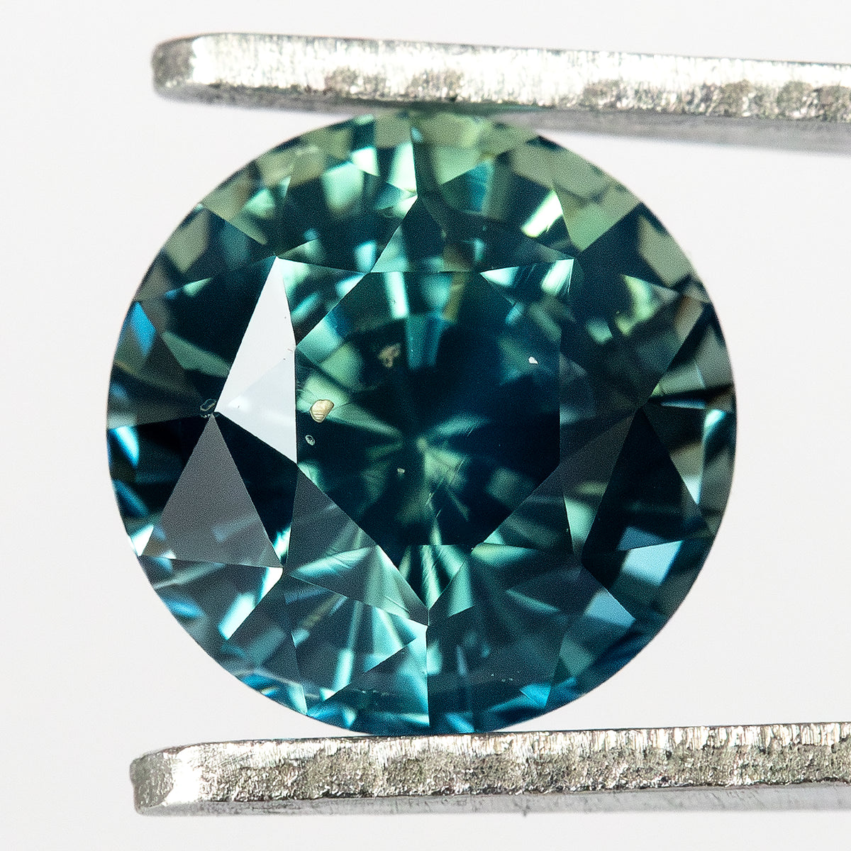 Round teal-blue and green sapphire face-up in tweezers, with dark central reflections and bright blue-green facet flashes.