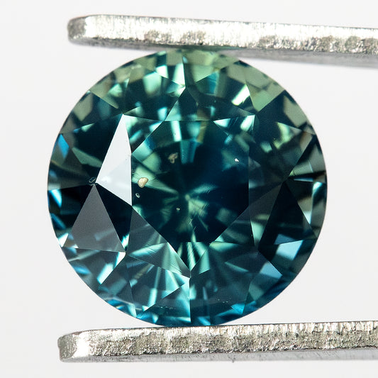 Round teal-blue and green sapphire face-up in tweezers, with dark central reflections and bright blue-green facet flashes.