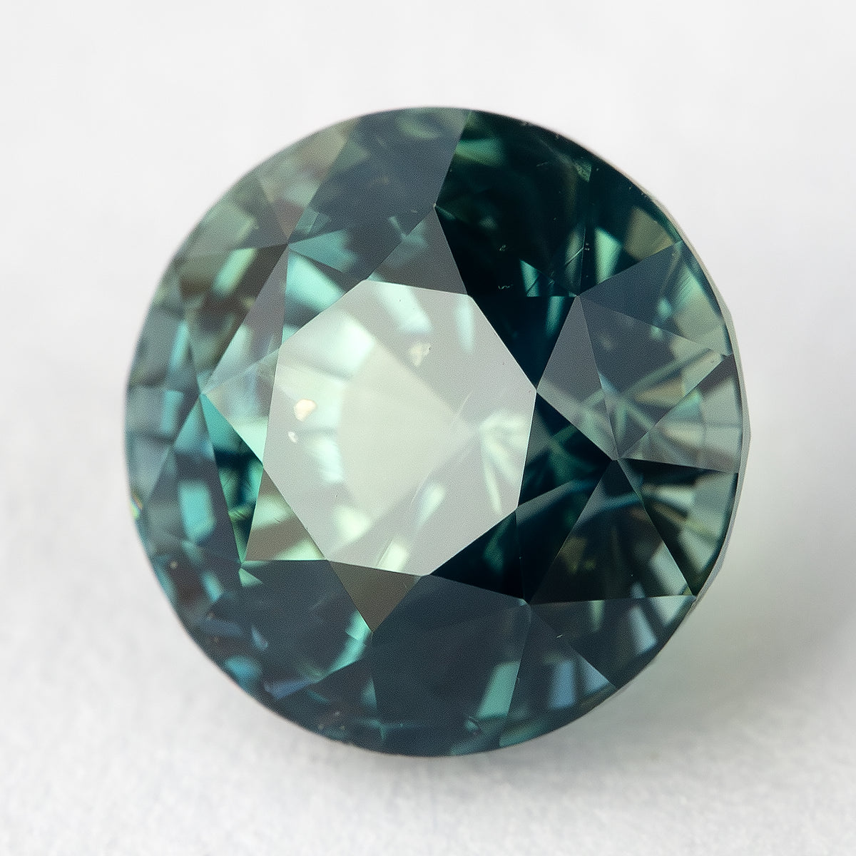Tilted face-up view of the loose round teal blue green sapphire resting on a light surface.