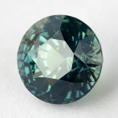 Tilted face-up view of the loose round teal blue green sapphire resting on a light surface.