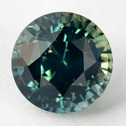 Round teal-blue and green sapphire tilted face-up on a light surface, with a dark central table and pale edge reflections.