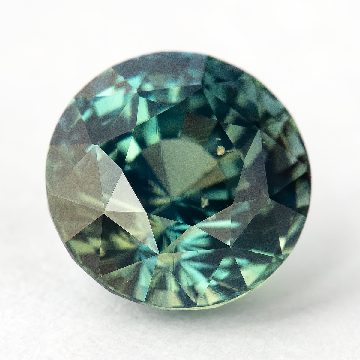 Round teal-blue and green sapphire tilted face-up on a light surface, with pale green and blue reflections across its facets.