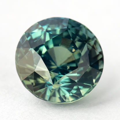 Round teal-blue and green sapphire tilted face-up on a light surface, with pale green and blue reflections across its facets.