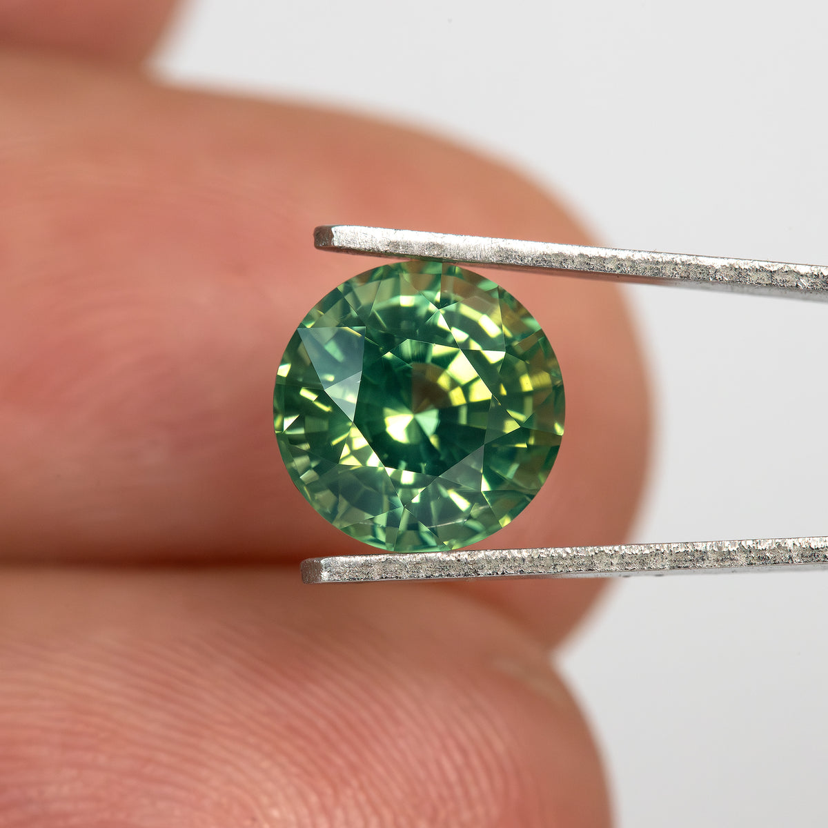 Round olive teal green sapphire held in tweezers between fingers to show scale.