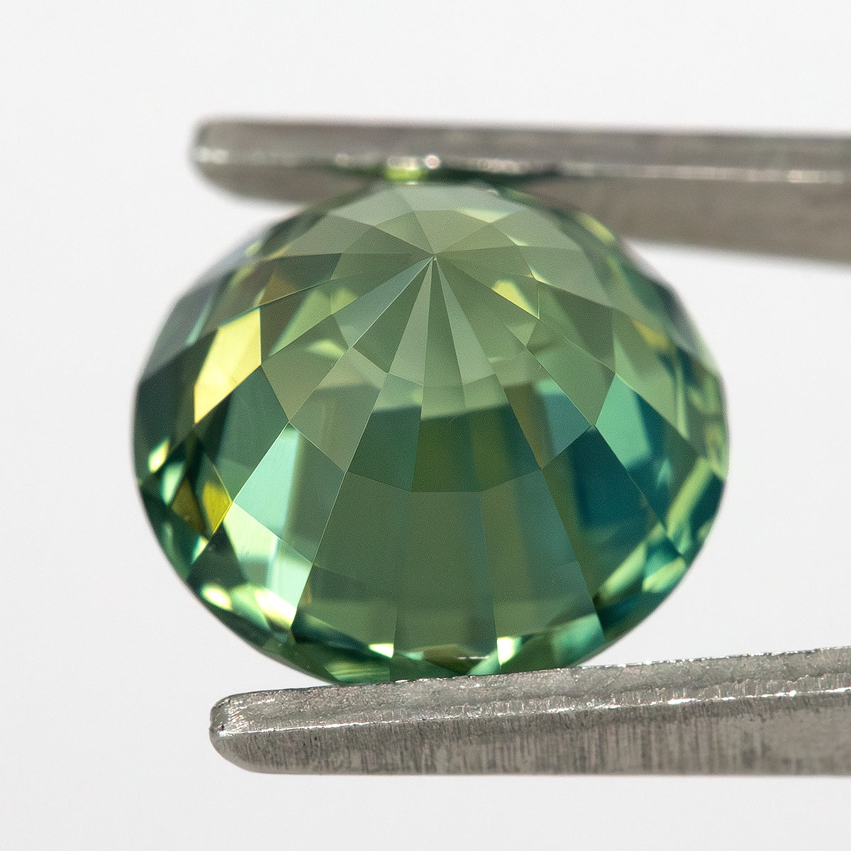 Oblique pavilion-side view of the round green sapphire held in tweezers, showing its pointed reverse and lower facets.