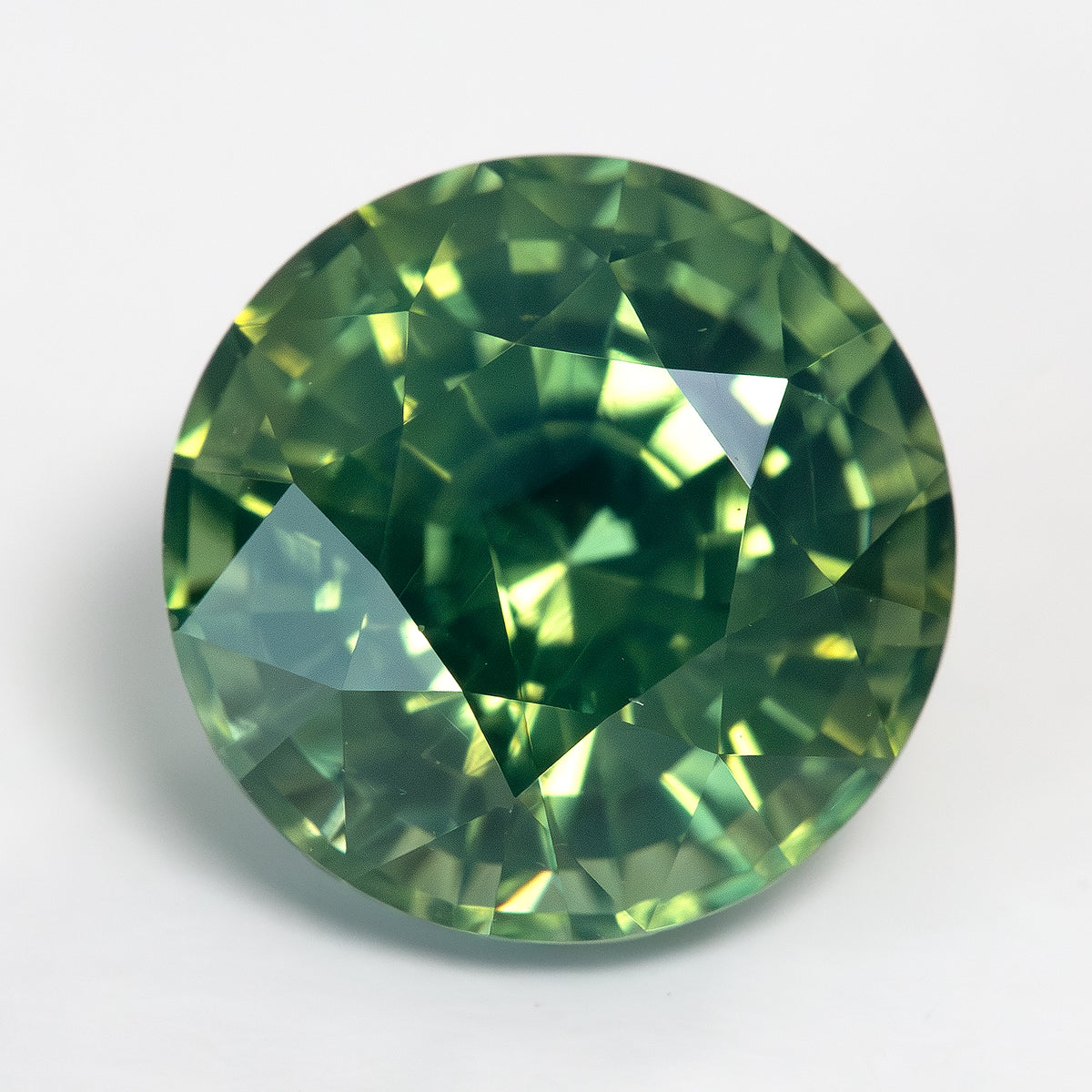 Round olive-green and teal sapphire face-up on a light surface, with dark central reflections and yellow-green flashes.