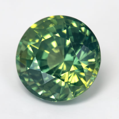 Tilted face-up view of the round olive-green and teal sapphire on a light surface, with bright yellow-green reflections.