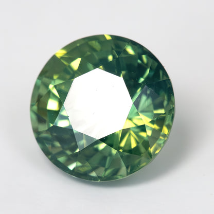 Round olive-green and teal sapphire face-up on a light surface, with a large white reflection across its table.