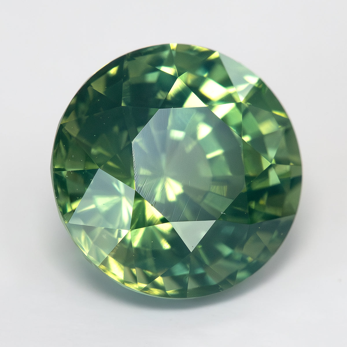 Round olive-green and teal sapphire tilted face-up on a light surface, with a pale reflection over the central table.