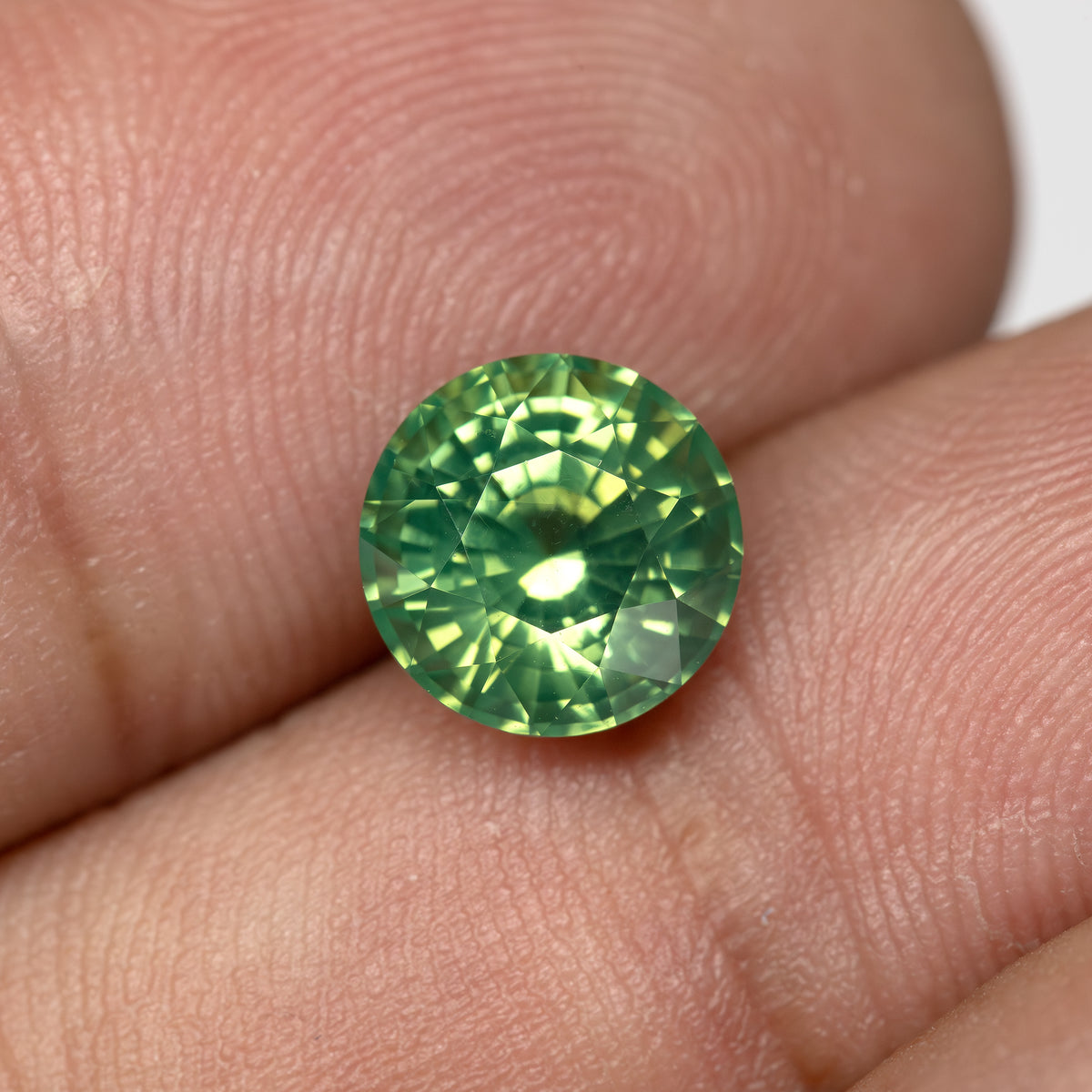 Close-up scale view of a round olive teal green sapphire held between fingers, alternative angle.