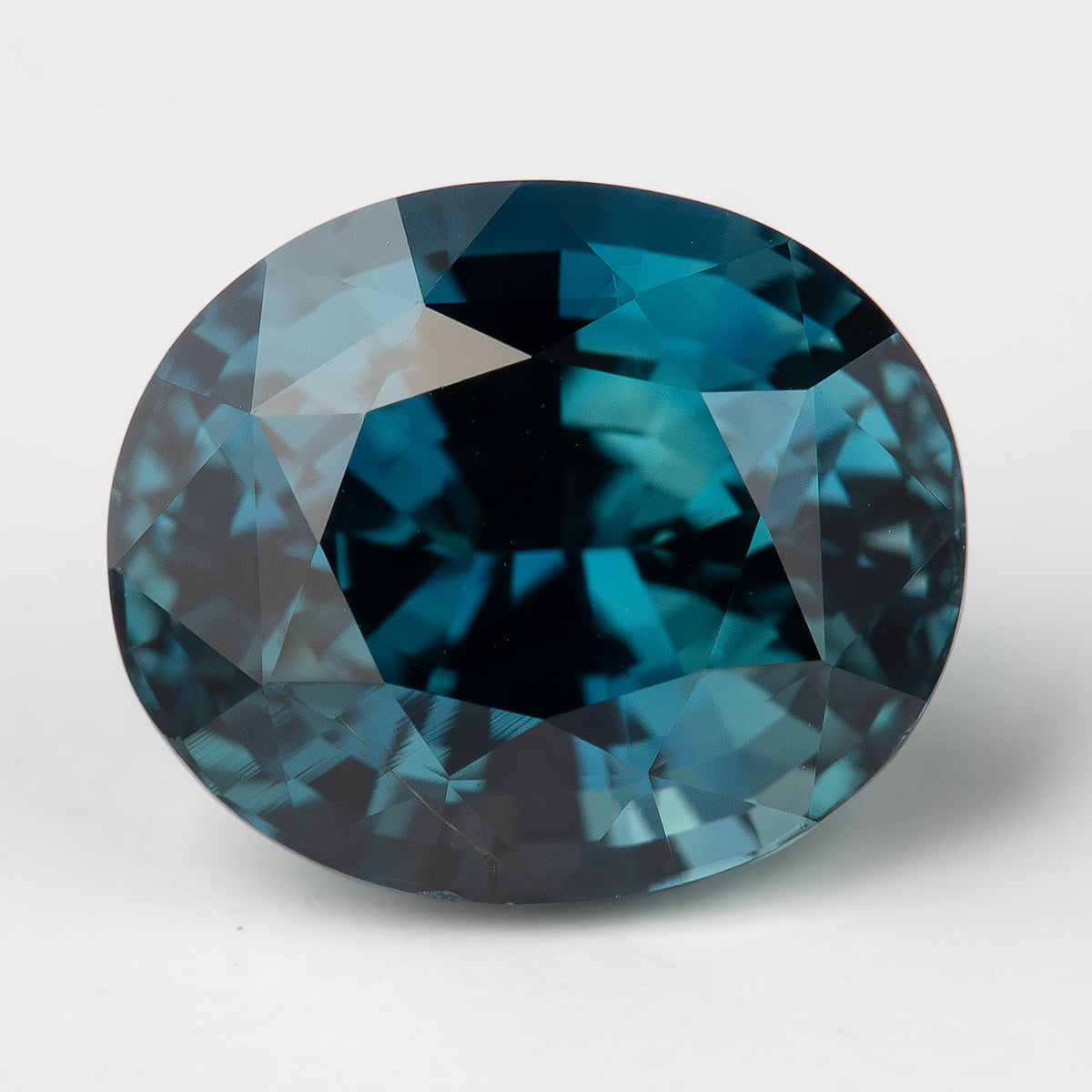 Face-up oval teal-blue sapphire on a white surface, with a dark central reflection and blue-green crown facets.