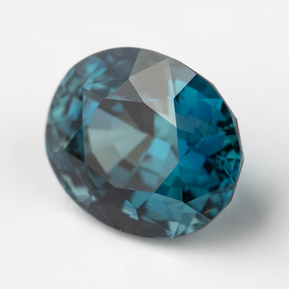 Oval teal-blue sapphire tilted on white, with pale reflections across the table.