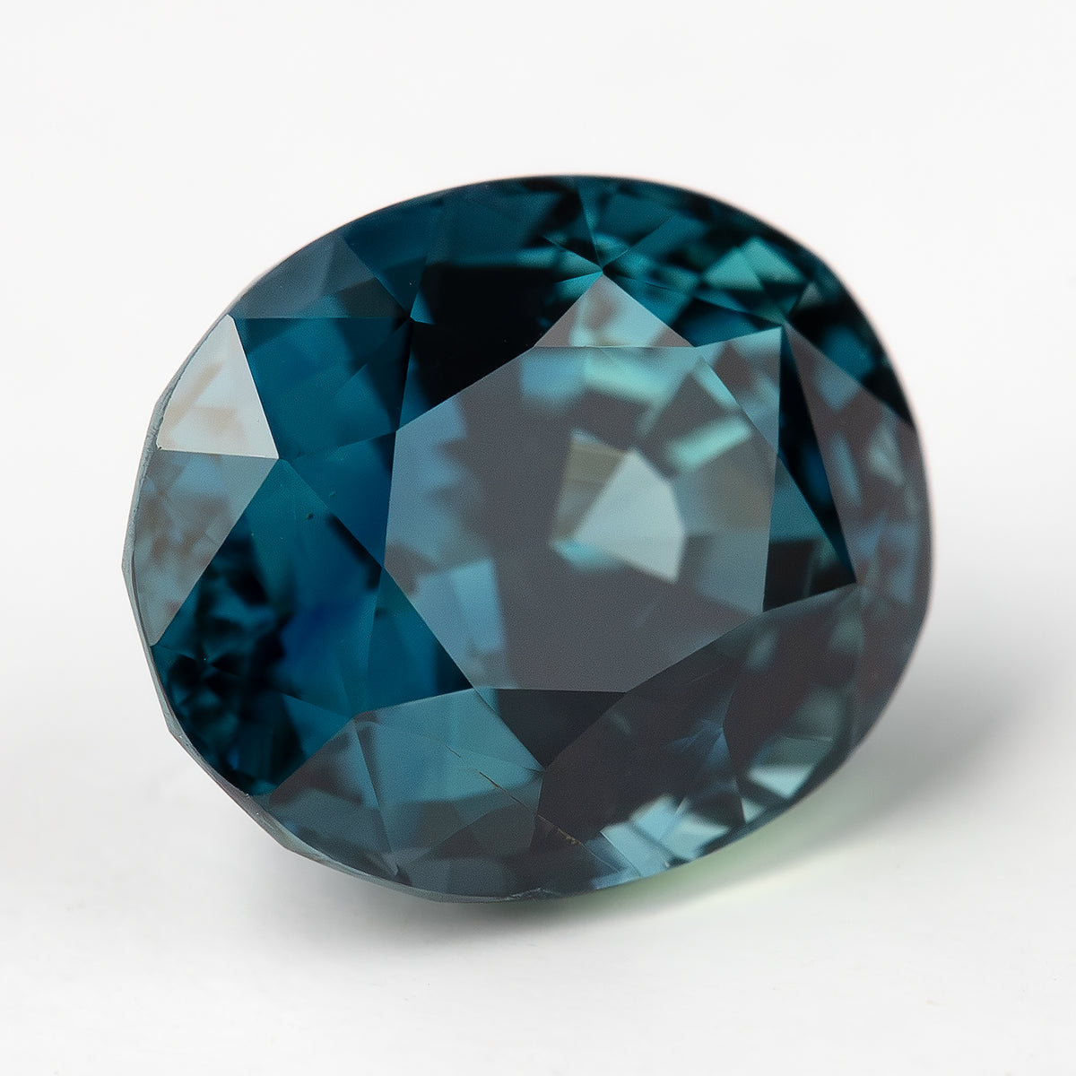 Oval teal-blue sapphire angled face-up on white, showing the table and dark-to-blue facet reflections.