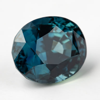 Oval teal-blue sapphire angled face-up on white, showing the table and dark-to-blue facet reflections.