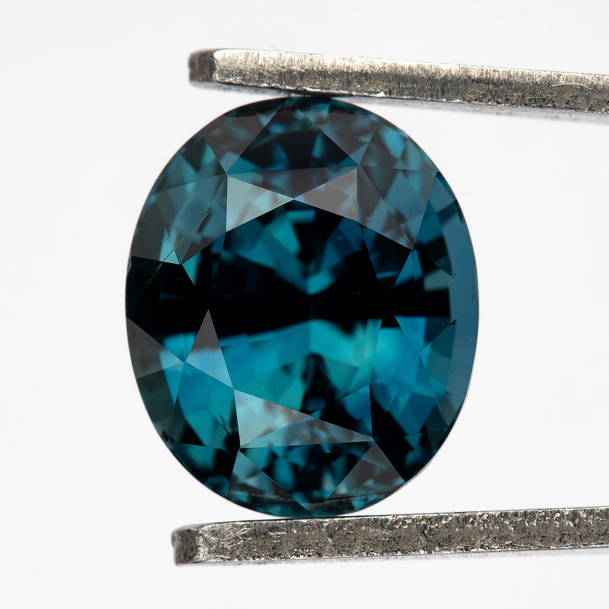 Teal-blue oval sapphire held face-up vertically in tweezers against a white background.