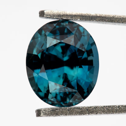 Teal-blue oval sapphire held face-up vertically in tweezers against a white background.