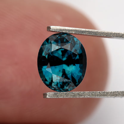 Teal-blue oval sapphire held face-up in tweezers against a fingertip for scale.