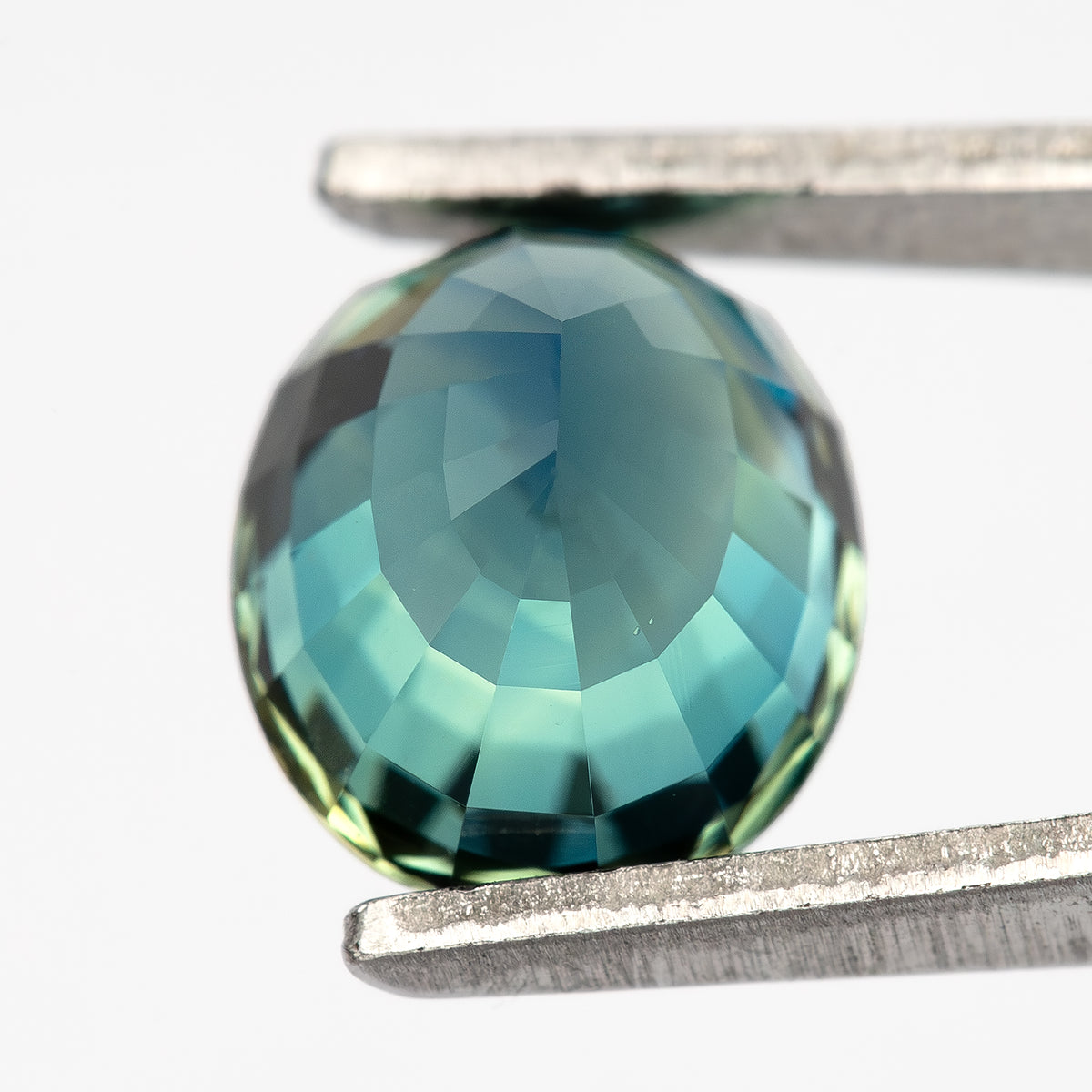 Angled pavilion view of the oval sapphire held in tweezers with pale reflections across the facets.