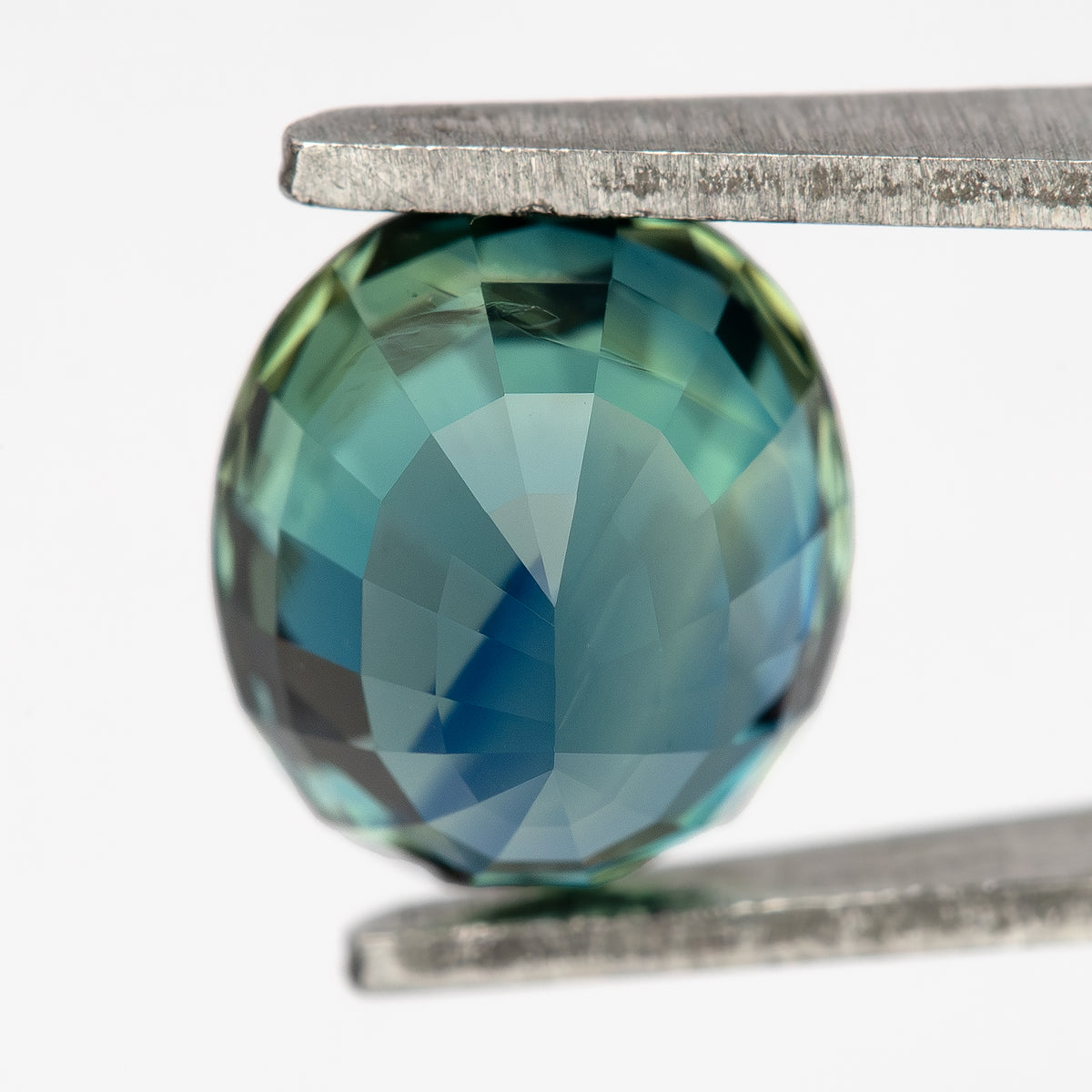Oval sapphire tilted pavilion-side toward the camera in tweezers, showing green-blue facet reflections.