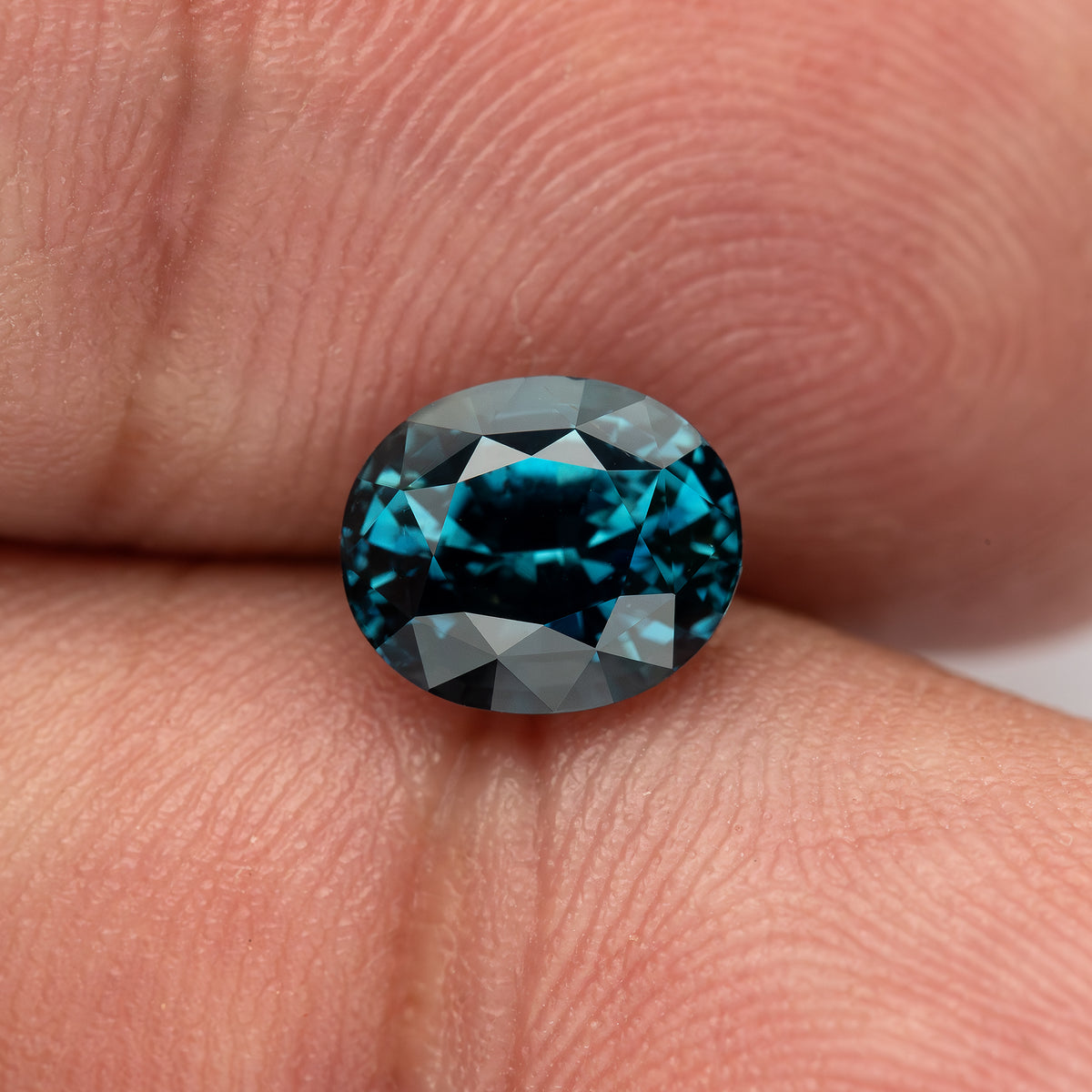 Oval teal-blue sapphire resting face-up between fingertips, with darker central reflections.