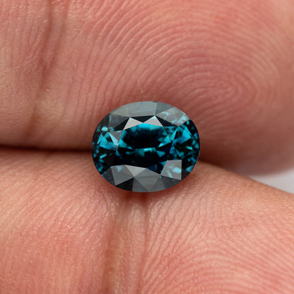 Oval teal-blue sapphire resting face-up between fingertips, with darker central reflections.