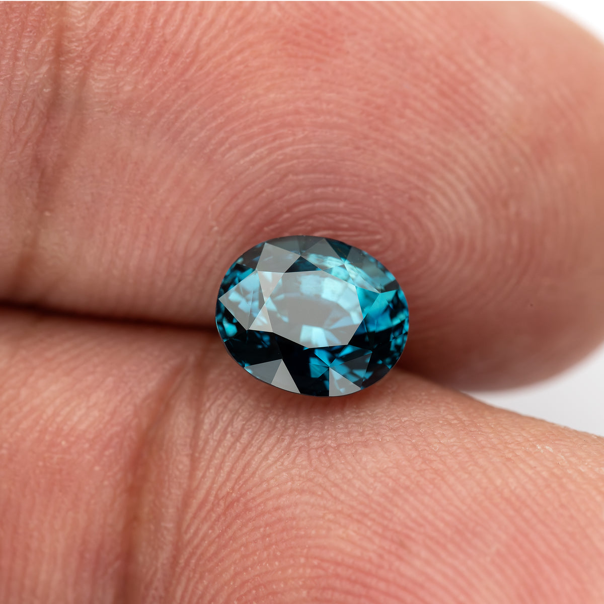 Oval sapphire tilted face-up between fingertips, with light across the table and blue-green edges.