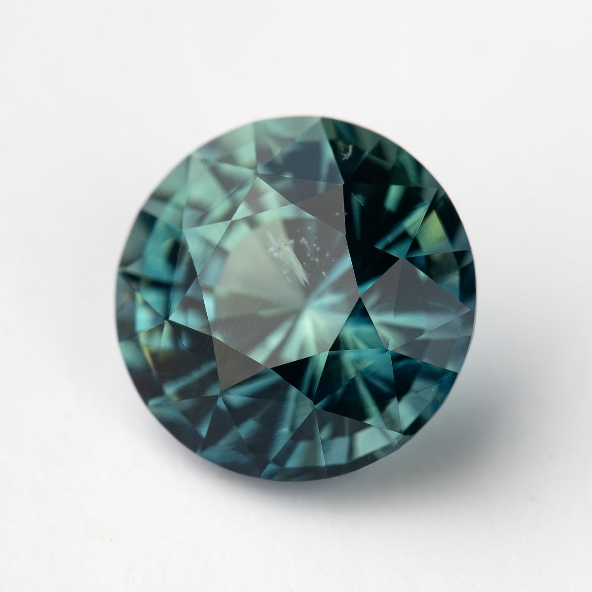 Round, deep teal blue-green sapphire viewed face up on a white background.