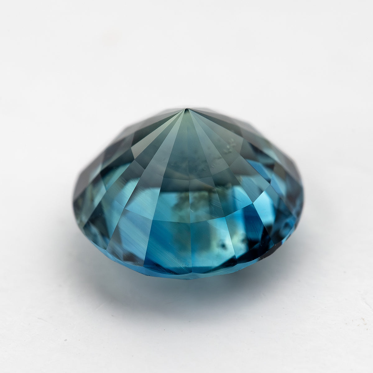 Round deep teal blue-green sapphire shown from a side angle on a light surface.