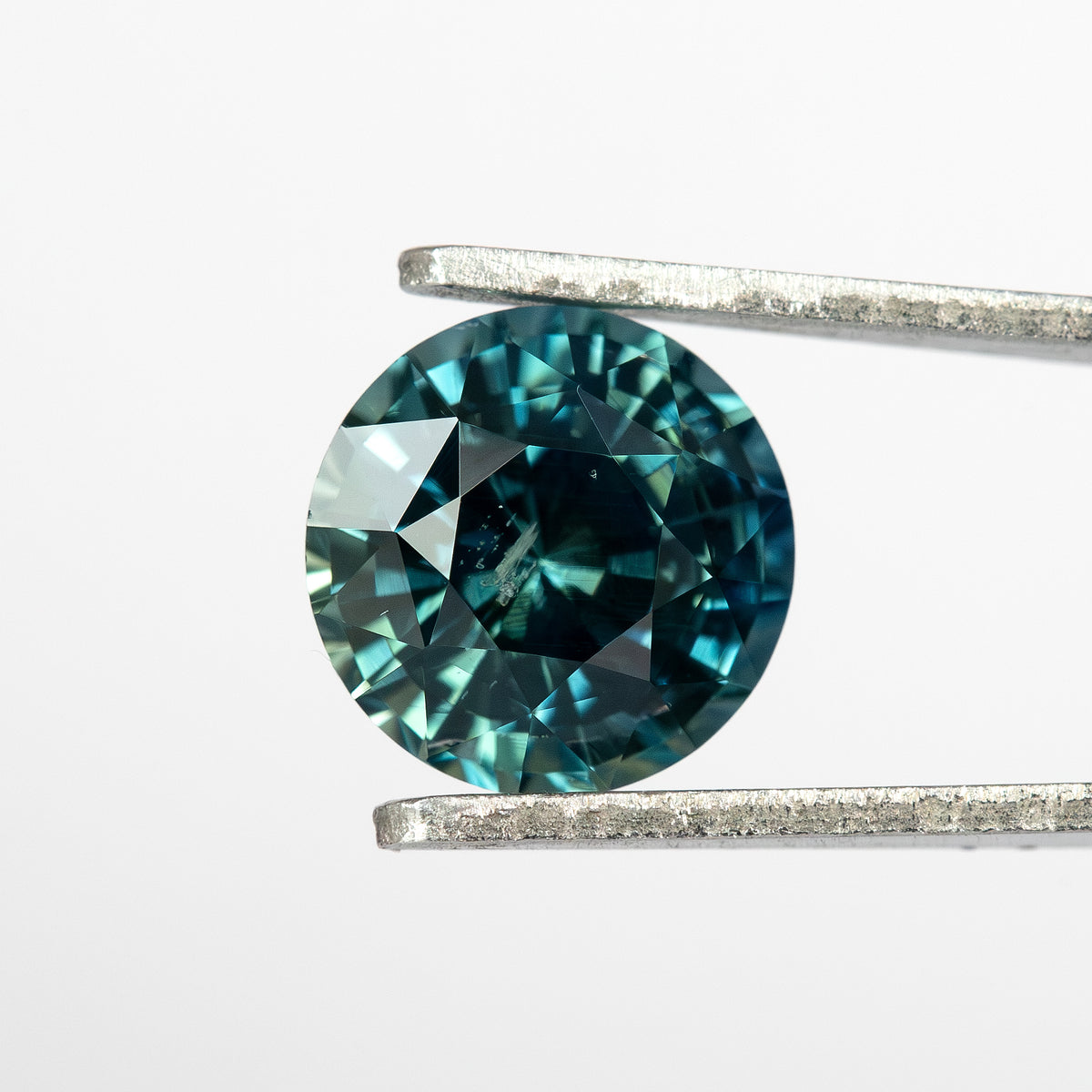 Round deep teal blue-green sapphire displayed face-up between tweezers against a plain background.