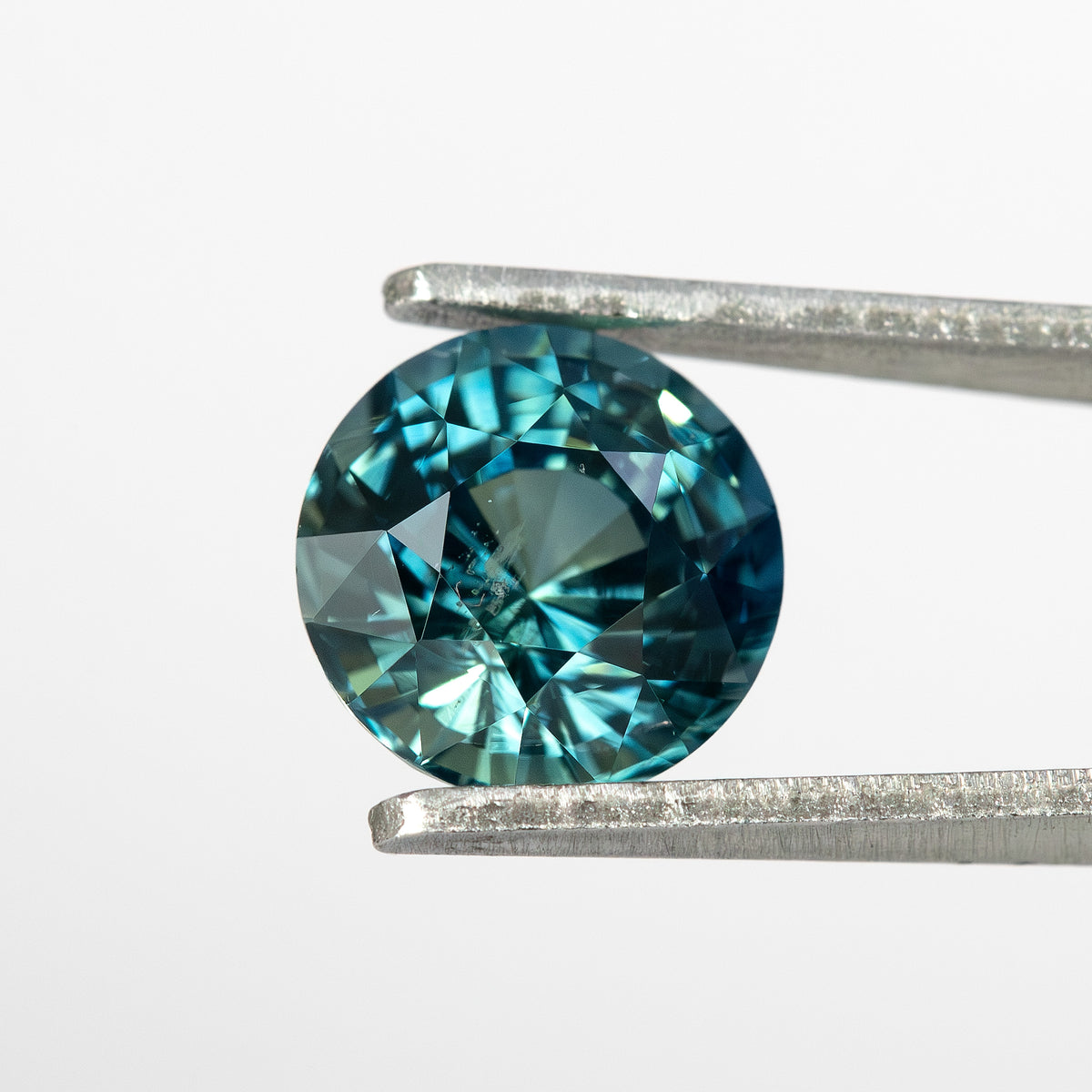 Round deep teal blue green natural sapphire held in tweezers, showing its step facets from a face-up angle.
