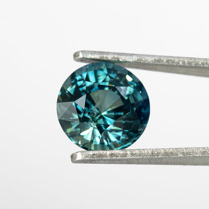 Round deep teal blue green natural sapphire held in tweezers, showing its step facets from a face-up angle.