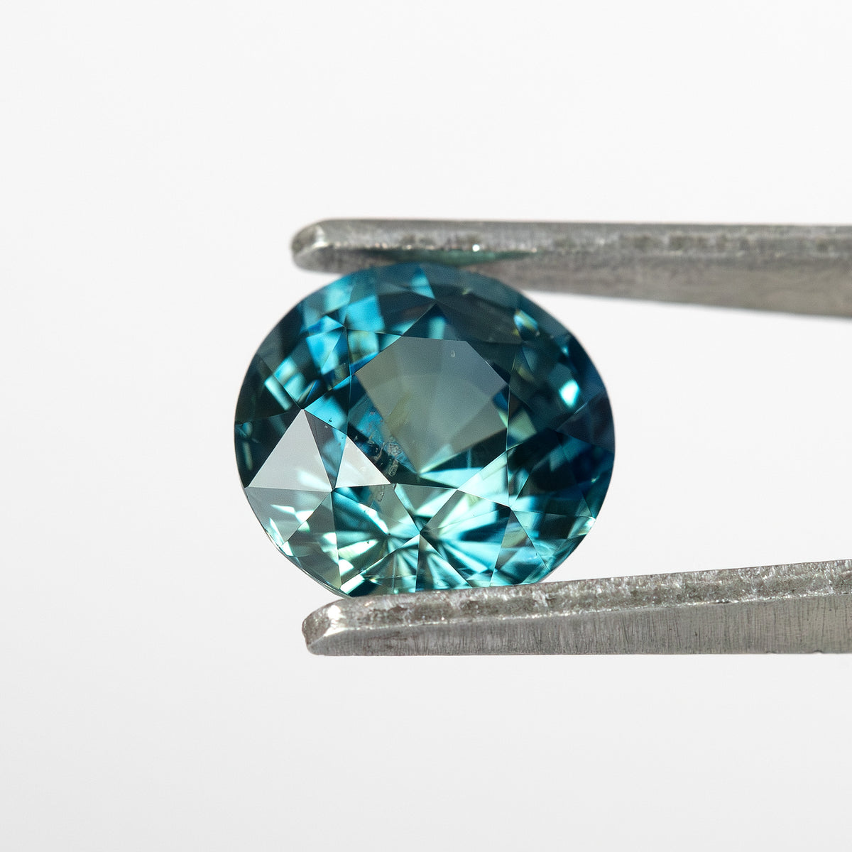 Round deep teal sapphire held in tweezers, showcasing its blue-green hues from a side angle.