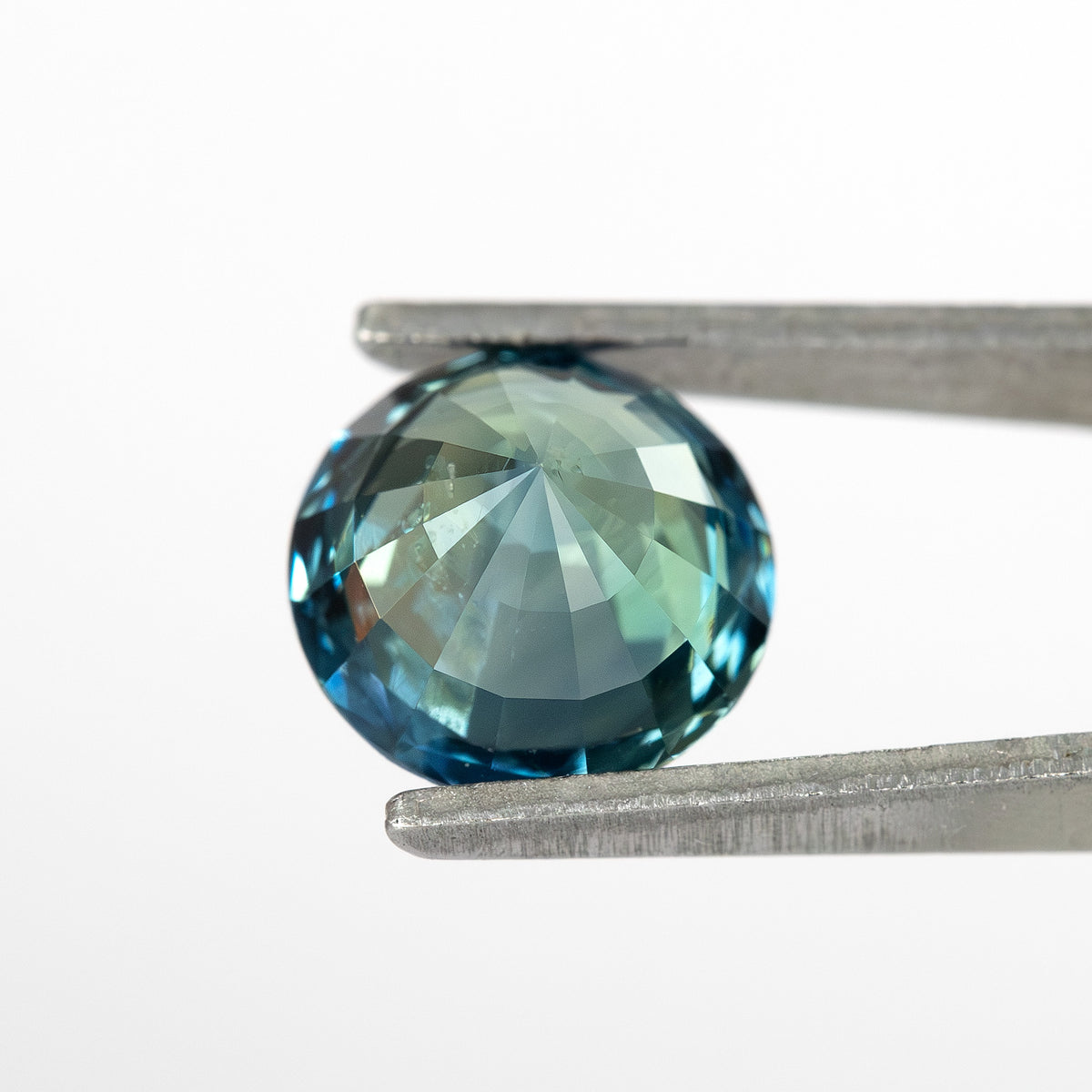 Round deep teal blue-green sapphire held in tweezers, viewed from the side, displaying faceted brilliance.