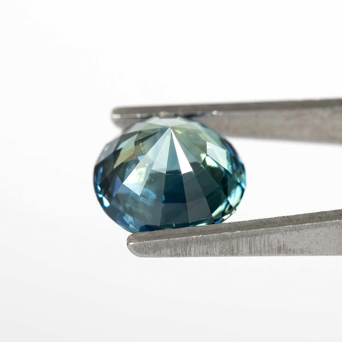 Round deep teal blue-green sapphire viewed from the side, held by tweezers against a white background.